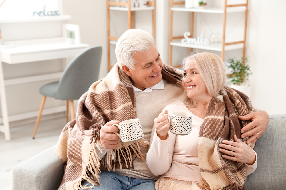 Happy older couple wrapped in blankets drinking coffee on couch in comfortable remodeled home – Home Remodeling Consulting