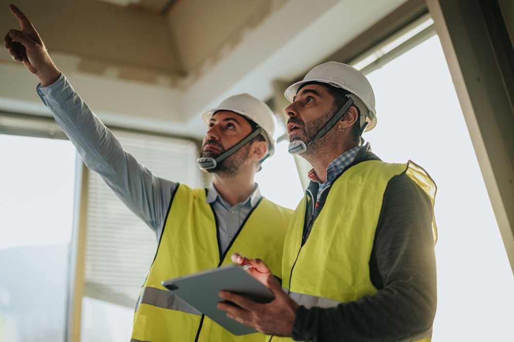 Construction Experts Inspecting Site and Reviewing Project Details – New Construction Guidance Two builders wearing hard hats and safety vests evaluating interior space for new construction guidance – New Construction Guidance