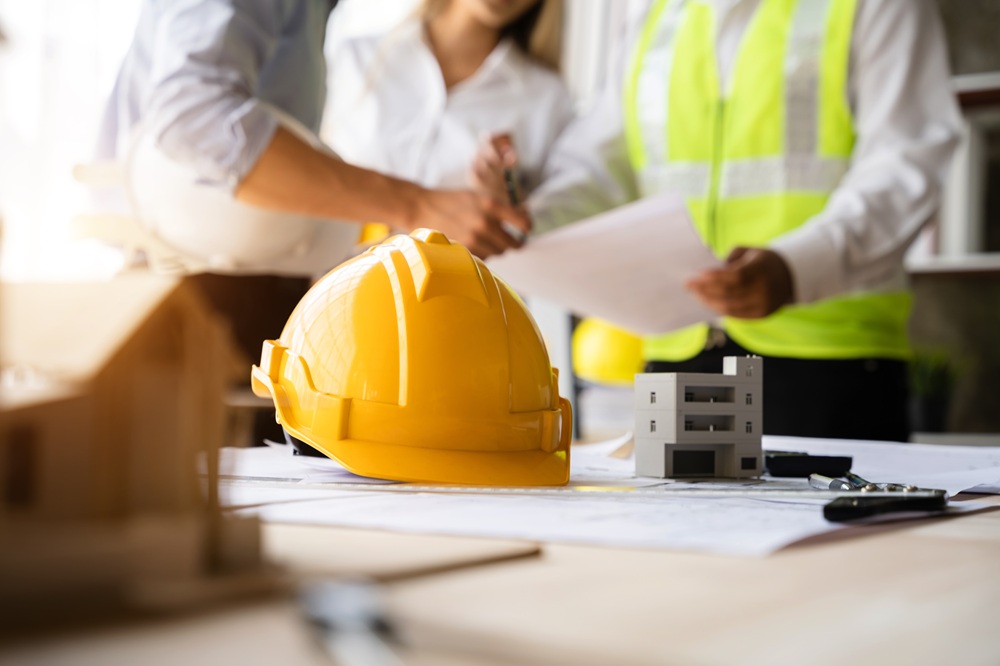 Construction Team Reviewing Building Plans and Safety Equipment – New Construction Guidance Hard hat and model home on table while professionals discuss blueprints for project planning – New Construction Guidance